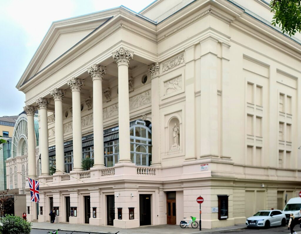 Bow Street facade of the Royal Opera House in London, with its Corinthian portico and the glass vault of the Floral Hall on the left.